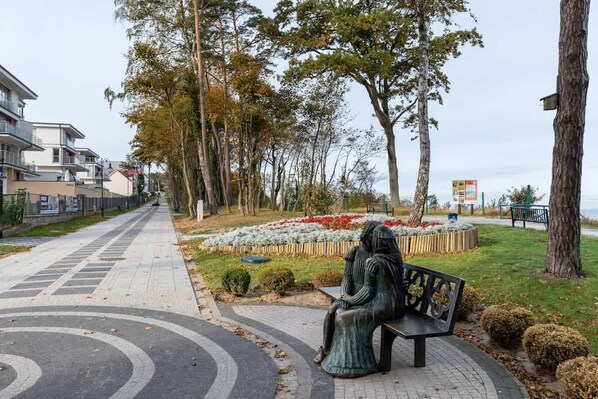 A park with a statue of a woman sitting on a bench, surrounded by trees and flower beds.