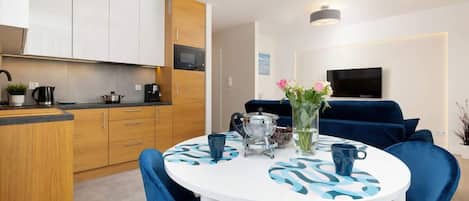 A modern kitchen with wooden cabinets, a dining table with blue chairs, and a TV in the background.