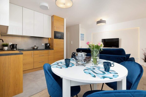 A modern kitchen with wooden cabinets, a dining table with blue chairs, and a TV in the background.
