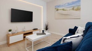 A modern living room with a flat-screen TV mounted on the wall, a wooden console table, a white coffee table, and a blue sofa with nautical-themed cushions. - Apartment with Balcony | Pool, Gym, 100 m to the Beach (Rewal)