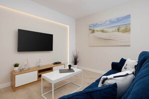 Una sala de estar moderna con un televisor de pantalla plana montado en la pared, una consola de madera, una mesa de centro blanca y un sofá azul con cojines de temática náutica.
