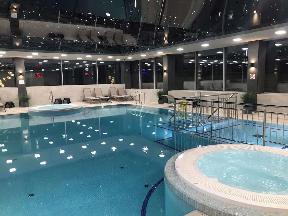 An indoor swimming pool with a hot tub, seating area, and a view of the night sky through the ceiling.