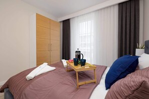 A hotel room with a bed, a wooden bedside table with a tray of breakfast items, a coffee maker, and a large window with curtains.