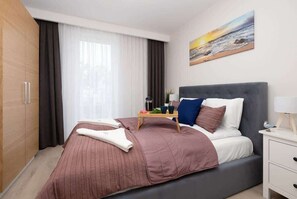 Una habitación de hotel moderna con una cama grande, una mesita de noche con lámpara, un cuadro con una escena de playa y un armario.