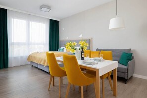 A modern hotel room with a bed, a dining area with yellow chairs, a grey sofa, and a white pendant light.