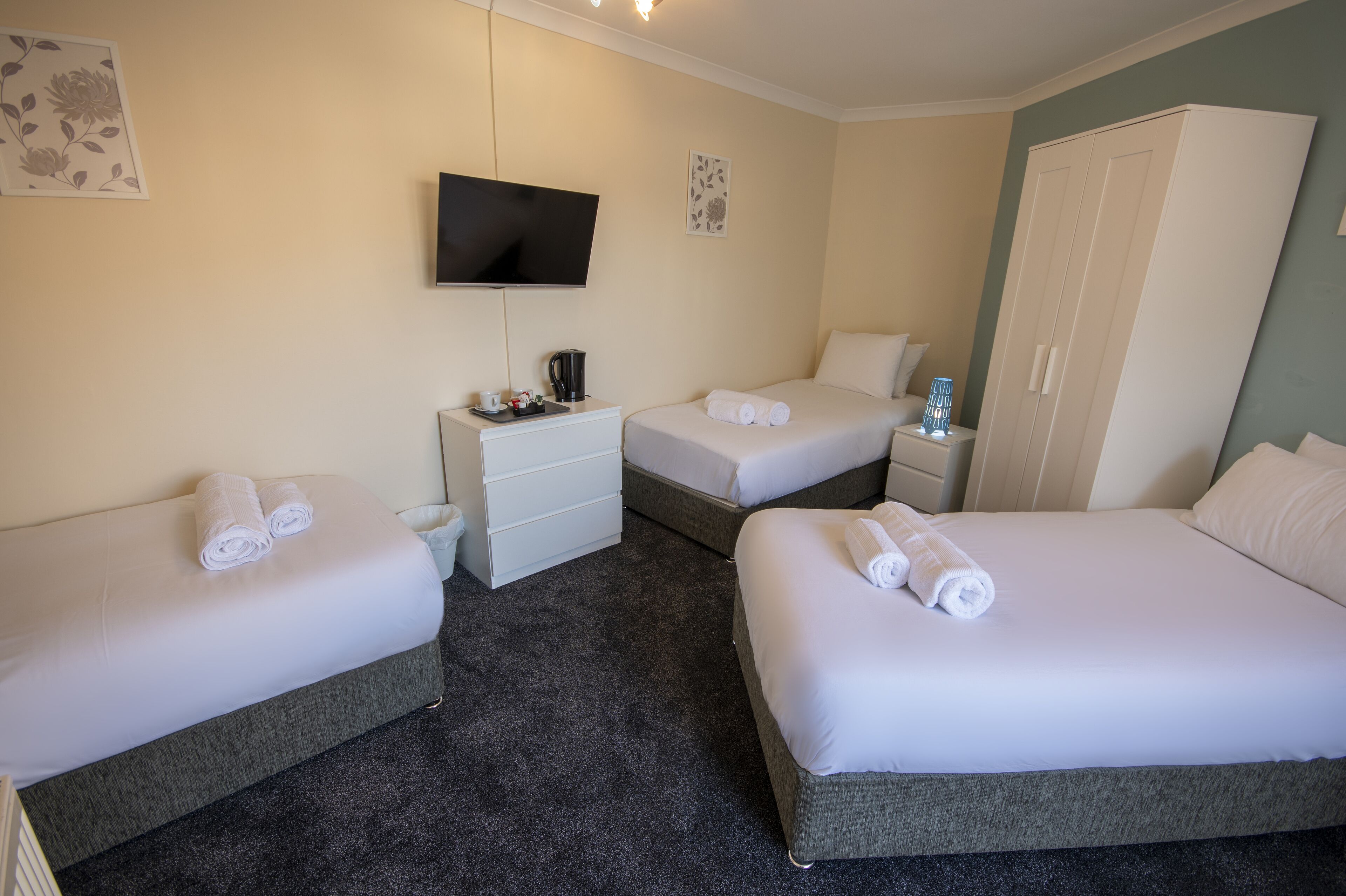 A hotel room with two single beds, a TV mounted on the wall, a wardrobe, and a small bedside table with a lamp.