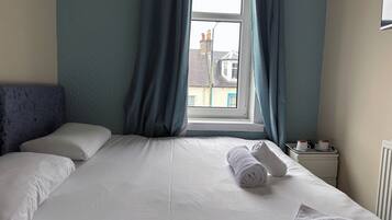 A hotel room with a bed, a window with curtains, a radiator, and a small table with cups.