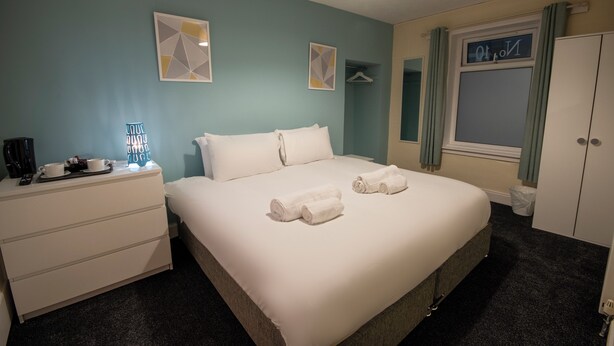 A neatly made bed with towels, a bedside table with a lamp, and a wardrobe. - Irvine Riverside Guesthouse (Irvine)