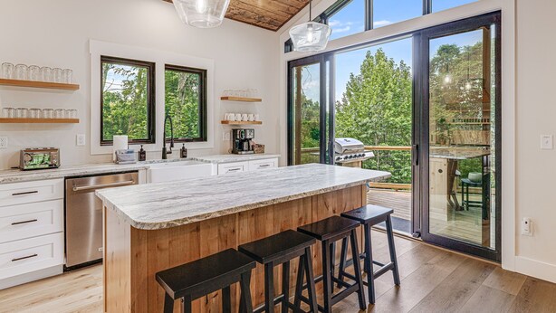 A modern kitchen with a large island, bar stools, and a sliding glass door leading to an outdoor area. - Pet-Friendly w/ Deck, Views + Biltmore Pass | Modern Maggie Valley Escape (Waynesville)