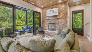 A spacious living area with a stone fireplace, large windows, and a comfortable seating arrangement. - Pet-Friendly w/ Deck, Views + Biltmore Pass | Modern Maggie Valley Escape (Waynesville)