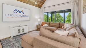A spacious living room with a large window, a beige sofa, a white cabinet, and a sign for Carolinamornings Luxury Vacation Rentals. - Pet-Friendly w/ Deck, Views + Biltmore Pass | Modern Maggie Valley Escape (Waynesville)