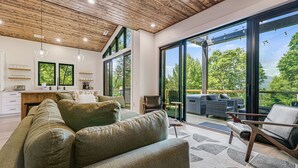 A modern living room with a large sofa, a dining area with a kitchen in the background, and a balcony with outdoor furniture. - Pet-Friendly w/ Deck, Views + Biltmore Pass | Modern Maggie Valley Escape (Waynesville)