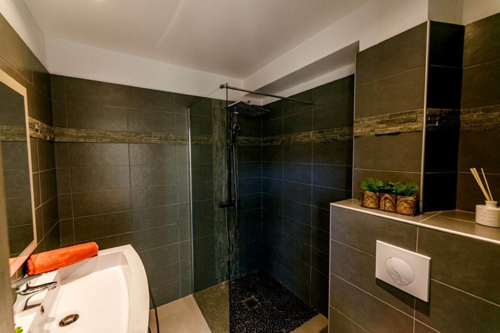 A modern bathroom with dark tiles, a glass shower enclosure, a white bathtub, and a small shelf with plants and toiletries.