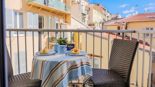 En balkon med et bord dækket til to, en fletstol og udsigt til omkringliggende bygninger.
