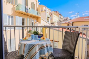 A balcony with a table set for two, a wicker chair, and a view of adjacent buildings. - SUEDE 3 AP4209 By Riviera Holiday Homes (Niza)