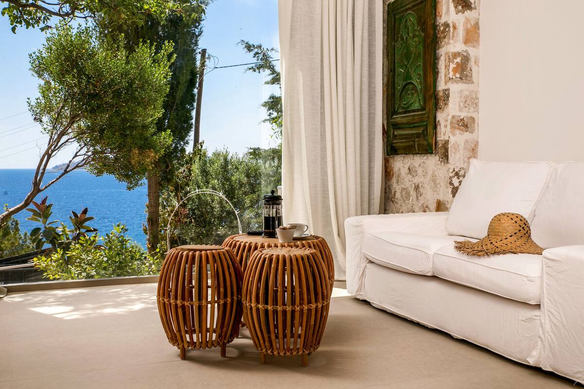 A room with a white sofa, a woven coffee table, and a view of the sea.
