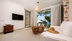 A modern living room with a flat-screen TV, a stone wall, a white sofa, wicker side tables, and a view of the sea through large windows. - Kale Suites (Kas)