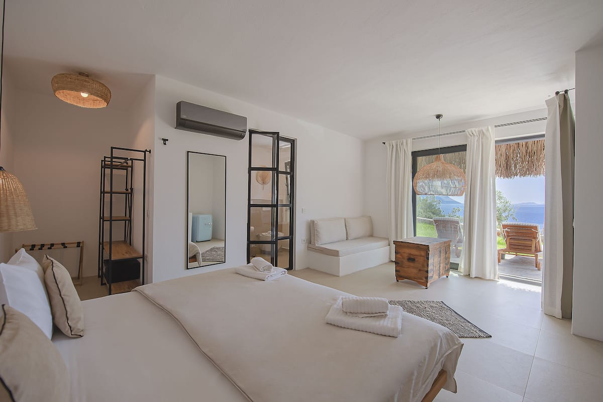 A spacious bedroom with a large bed, a wooden dresser, a seating area with a sofa and chair, and a balcony with a view.