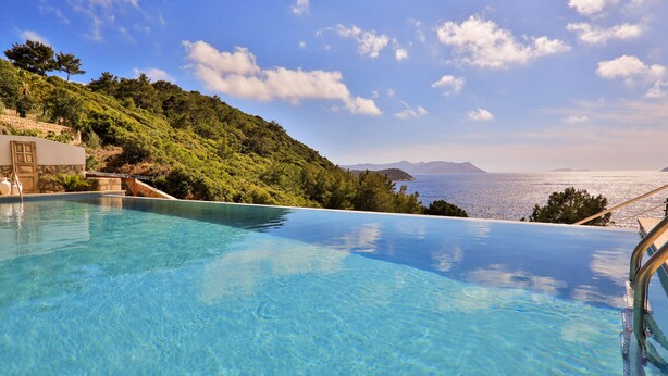 An infinity pool with clear blue water, surrounded by lush greenery and overlooking a scenic view of the sea and distant mountains. - Kale Suites (Kas)