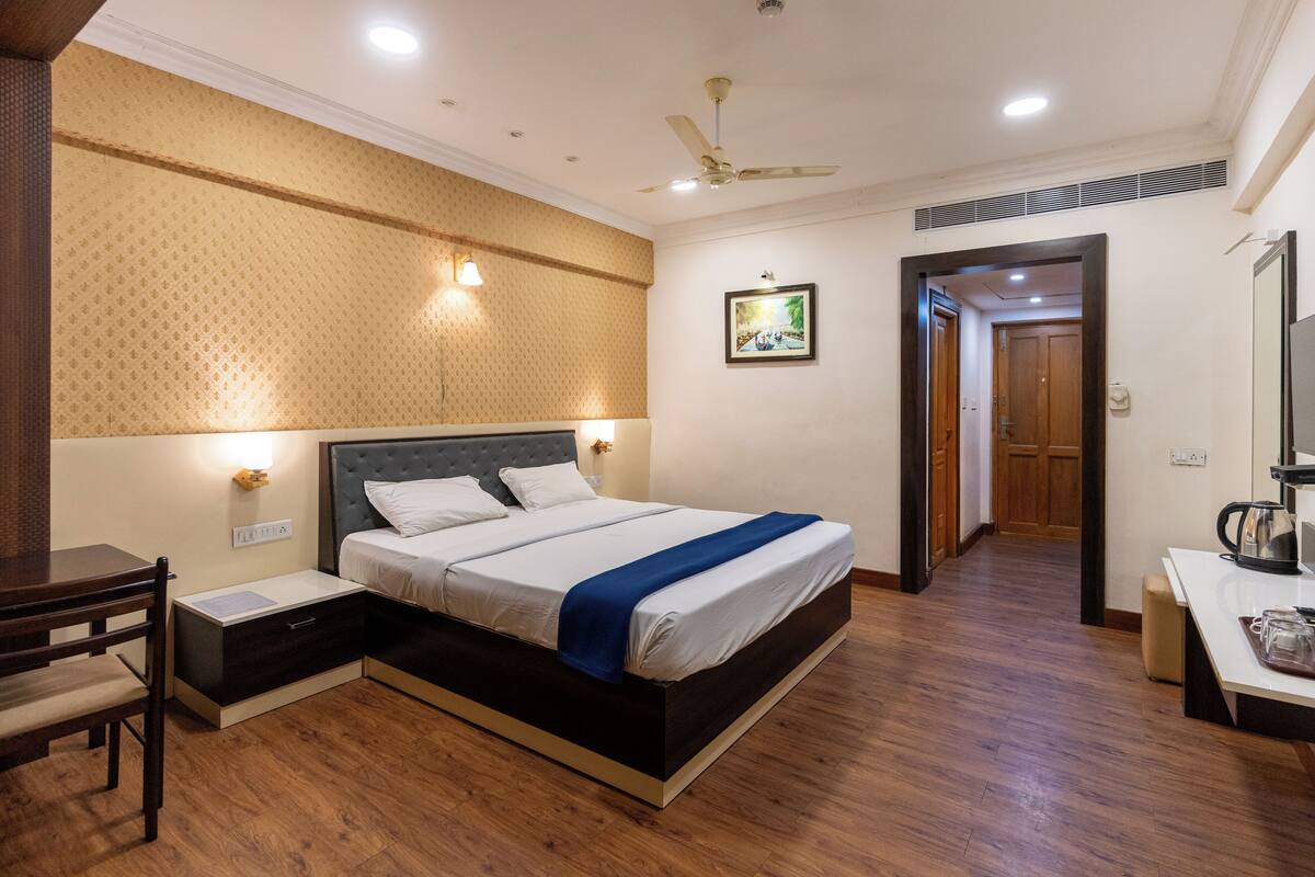 A hotel room with a bed, bedside table, chair, and a desk with a kettle.