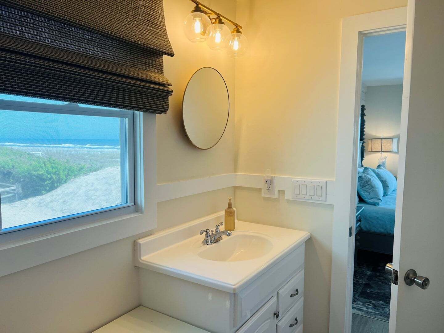 A bathroom with a window, a sink, a mirror, and a door leading to a bedroom.