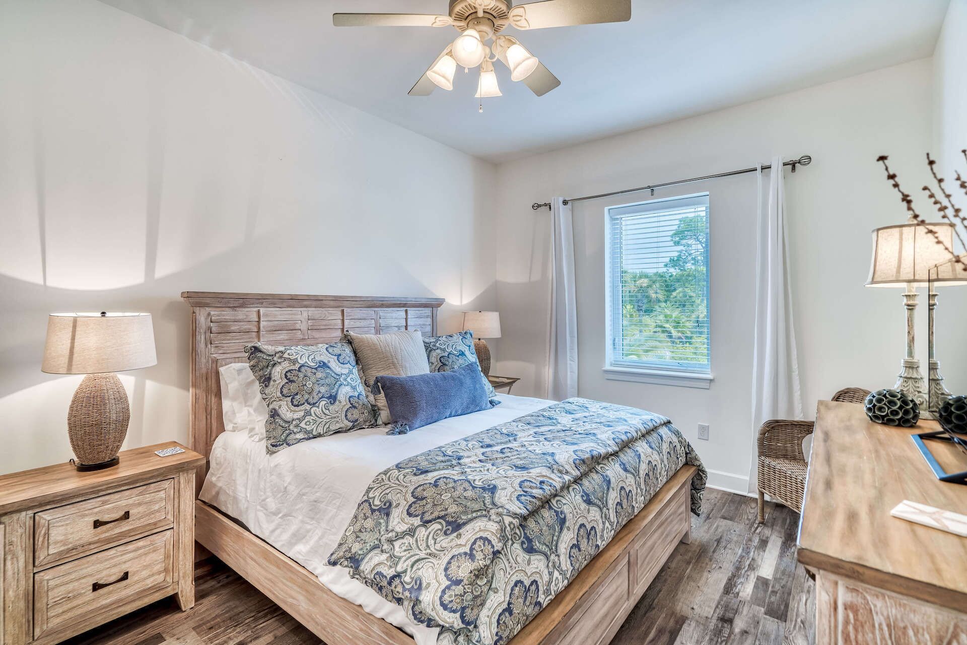 A bedroom with a wooden bed, patterned bedding, a nightstand with a lamp, a wooden dresser, and a ceiling fan.