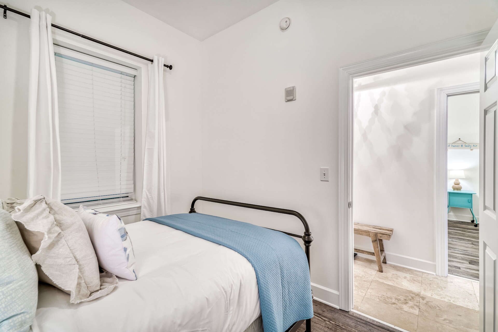 Une chambre à coucher avec un lit, une fenêtre avec des stores, une porte donnant sur une salle de bain et un petit tabouret en bois.