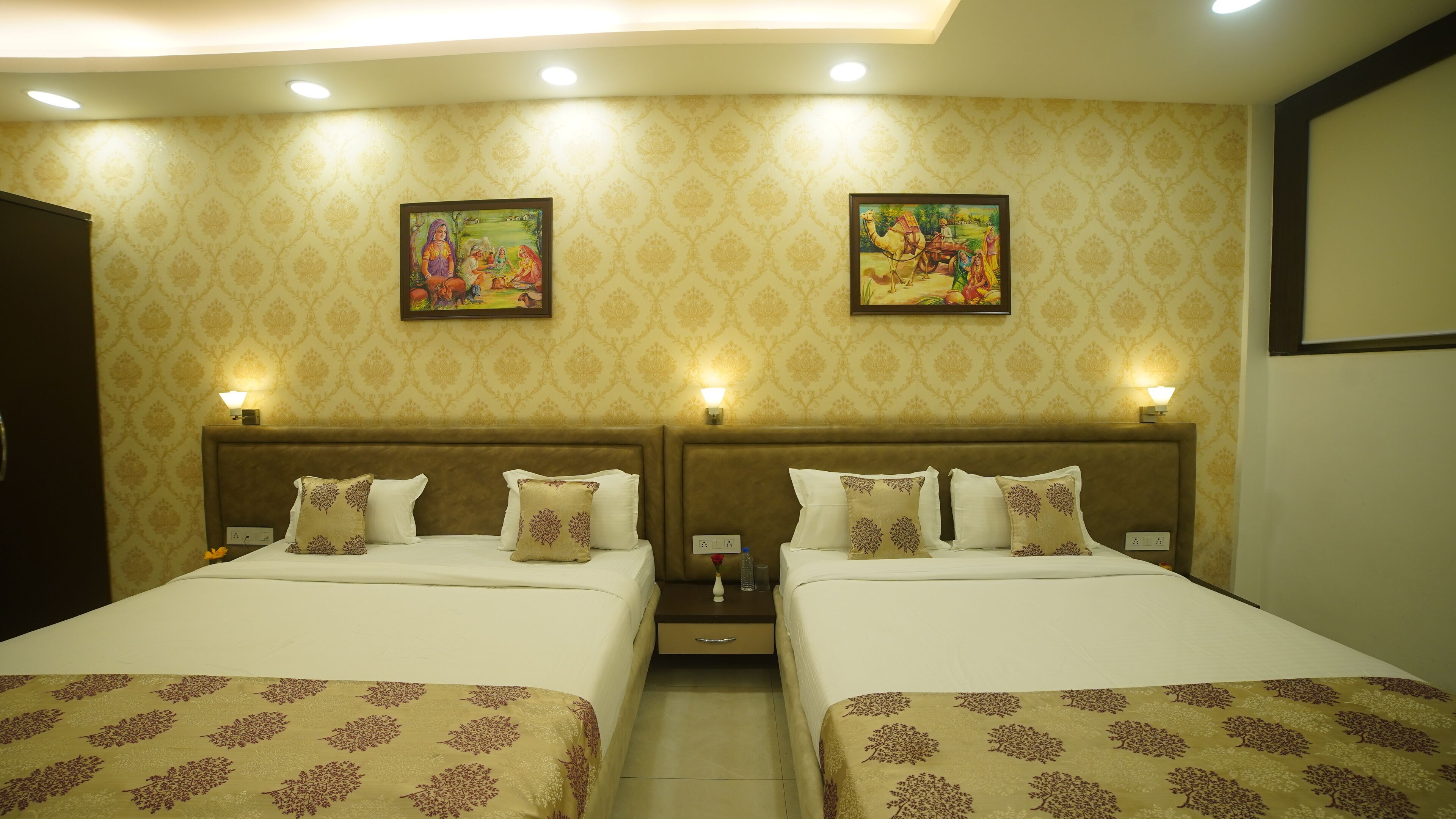 A hotel room with two beds, each with patterned bedspreads and matching pillows. There are two wall-mounted lamps and two framed paintings above the beds.