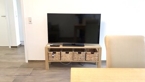 Una sala de estar con un televisor de pantalla plana sobre un soporte de madera, un control remoto y una tableta en el soporte. Hay cestas de mimbre debajo del mueble de la televisión y un sillón a la derecha.