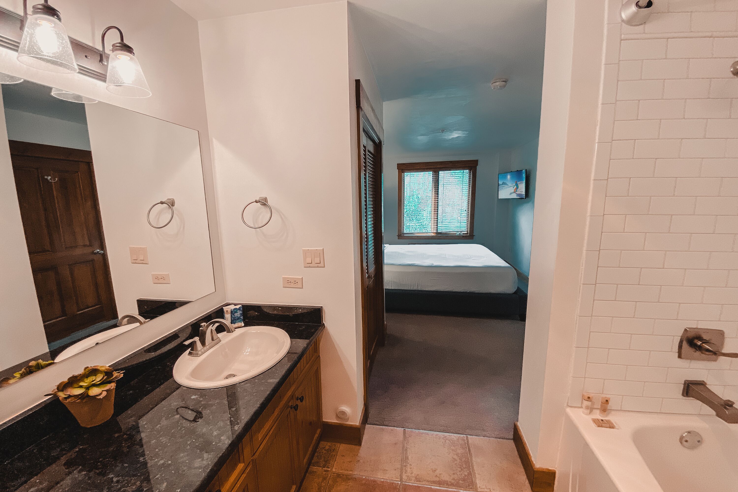 A bathroom with a sink, mirror, and shower. There is a window with blinds and a door leading to another room.