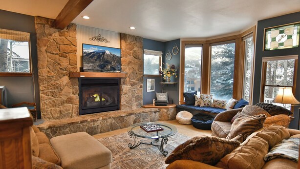 A cozy living room with a stone fireplace, comfortable seating, and large windows offering a view of the outdoors. - Ridge Retreat Breckenridge Townhome: Great Location, Views (Breckenridge)