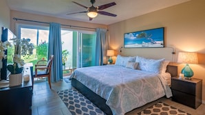 Une chambre à coucher avec un lit, un ventilateur de plafond, un tableau, une table de chevet, une lampe, une chaise, une table et une vue sur l’extérieur.