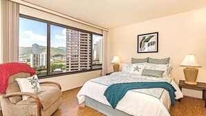 Hotellihuone, jossa on suuri ikkuna, sinisellä peitteellä varustettu sänky, punaisella heittohuovalla päällystetty tuoli ja kaksi yöpöytävalaisinta.