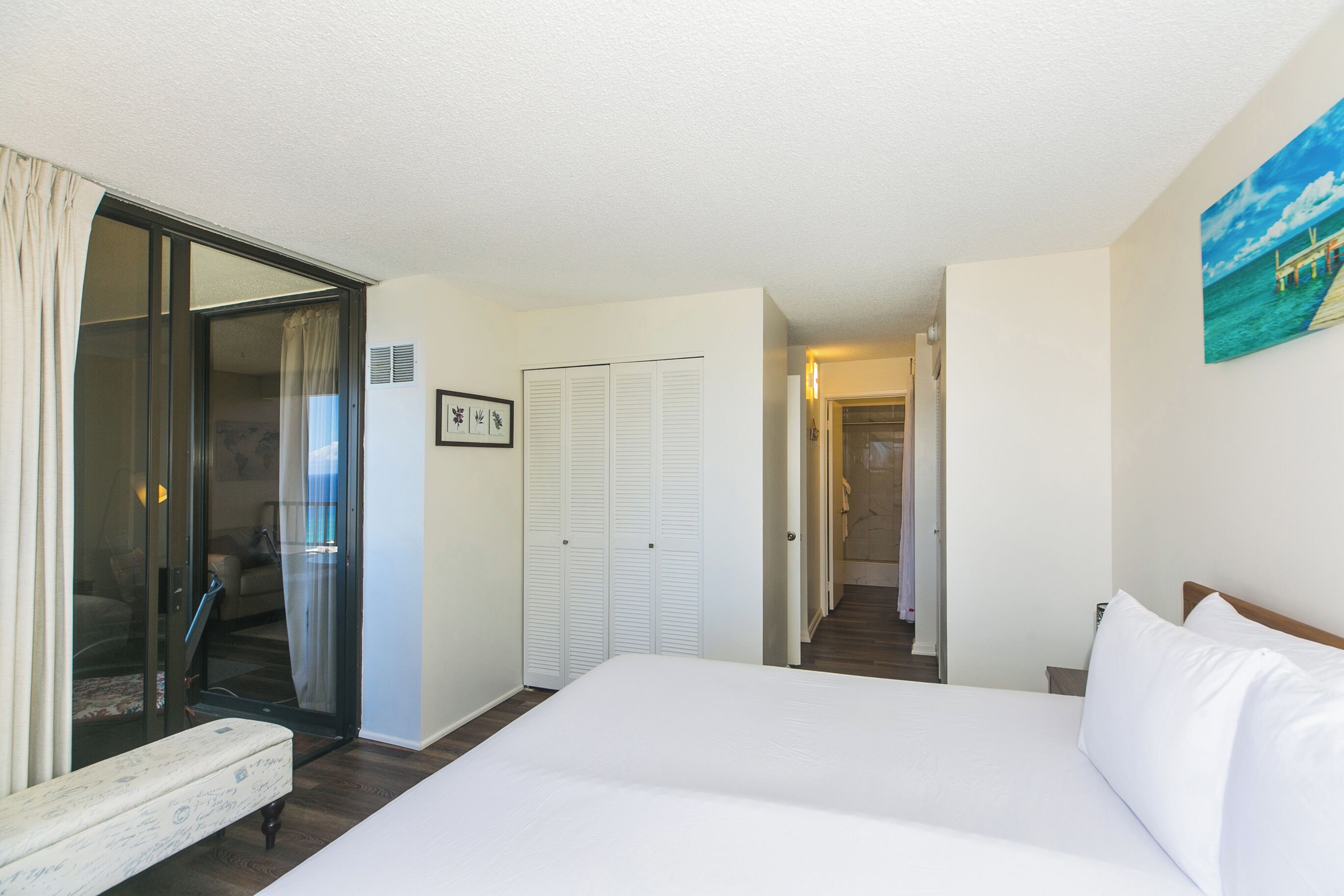 Une chambre d’hôtel avec un grand lit, un banc, une porte-fenêtre coulissante, un placard et un couloir menant à une autre pièce.