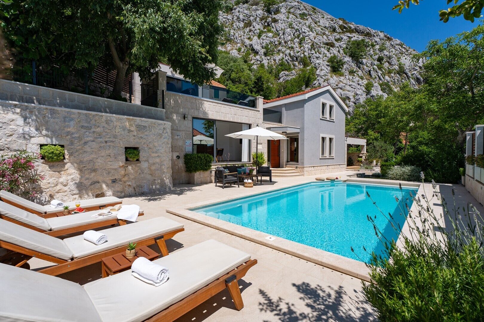 Ein Poolbereich mit Liegestühlen, ein kleines Schwimmbecken und ein Gebäude mit einer Steinmauer, im Hintergrund ein Berg.