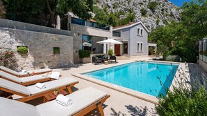 Area piscina con sedie a sdraio, una piccola piscina e un edificio con parete in pietra e montagna sullo sfondo.