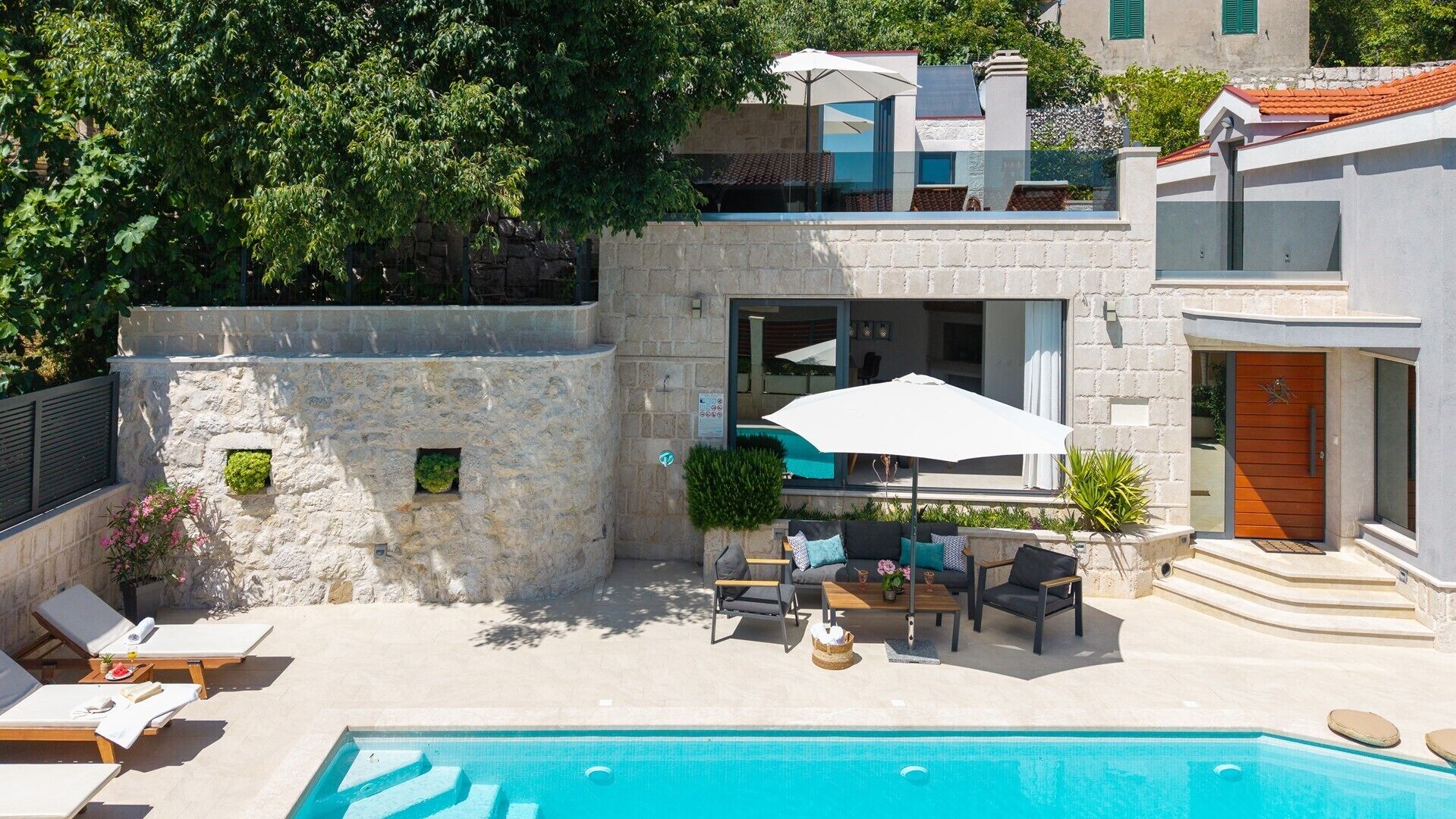Un espace piscine extérieur moderne, doté de chaises longues, d’une table avec des parasols et d’un mur en pierre.