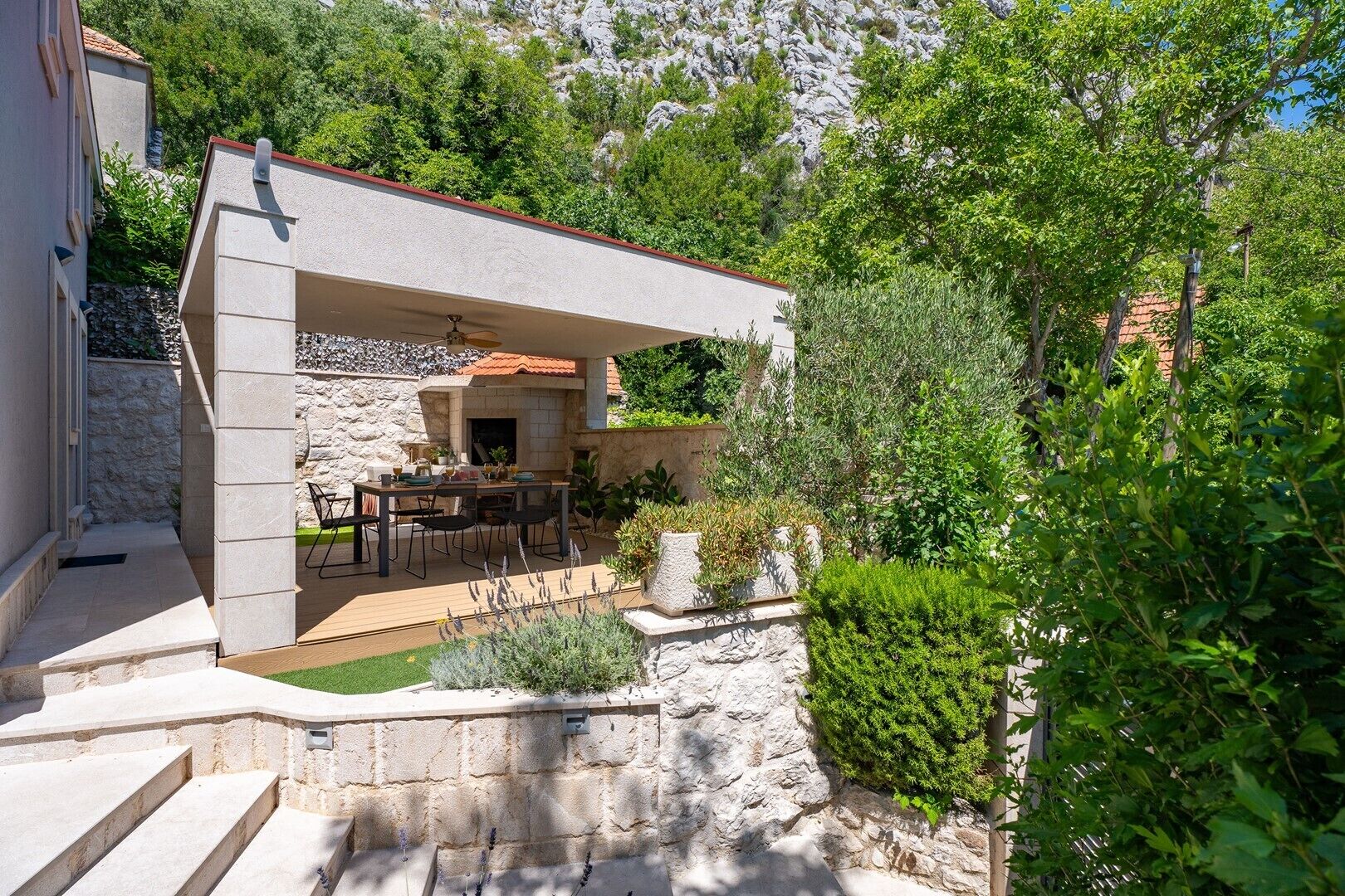 A stone patio with a table and chairs, surrounded by greenery and a stone wall.