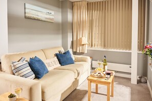 A living room with a beige sofa, a small wooden table with food and drinks, a lamp, and a window with curtains. - Bayfort Sea Studio (Torquay)
