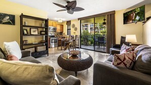 Sala de estar con sofá, sillones, una mesa de centro y una estantería. Hay un ventilador de techo, un televisor montado en la pared, y vistas a la cocina y al balcón.