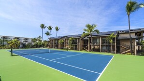 Una cancha de tenis con una red, rodeada de palmeras y un edificio con varios balcones.