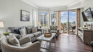 Ein geräumiges Wohnzimmer mit grauem Sofa, einem Korbstuhl, einem Glastisch und Blick ins Freie durch große Fenster.