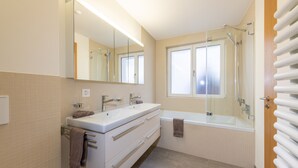 Une salle de bain moderne dotée d’un meuble-lavabo à deux vasques, d’un grand miroir, d’une baignoire et d’une fenêtre laissant entrer la lumière naturelle.