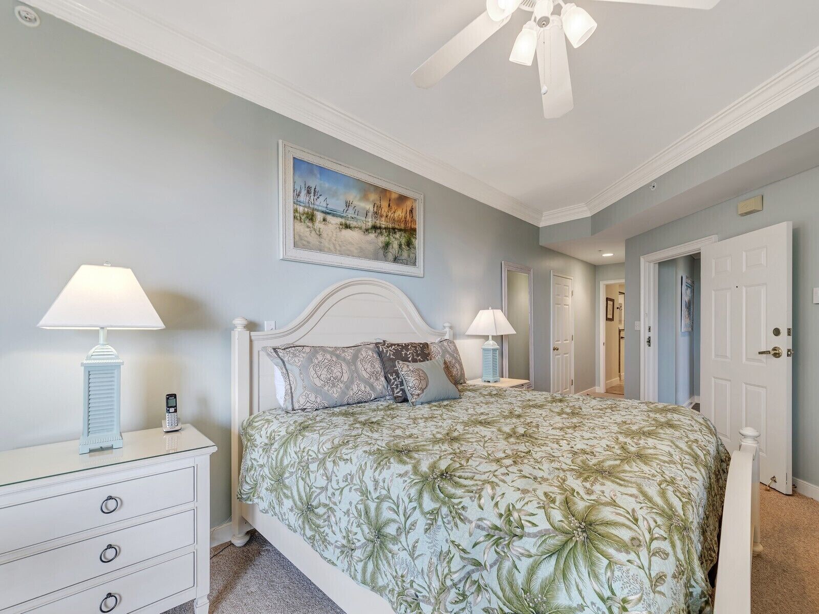 A bedroom with a bed, two bedside tables, a ceiling fan, a painting on the wall, and a door leading to another room.