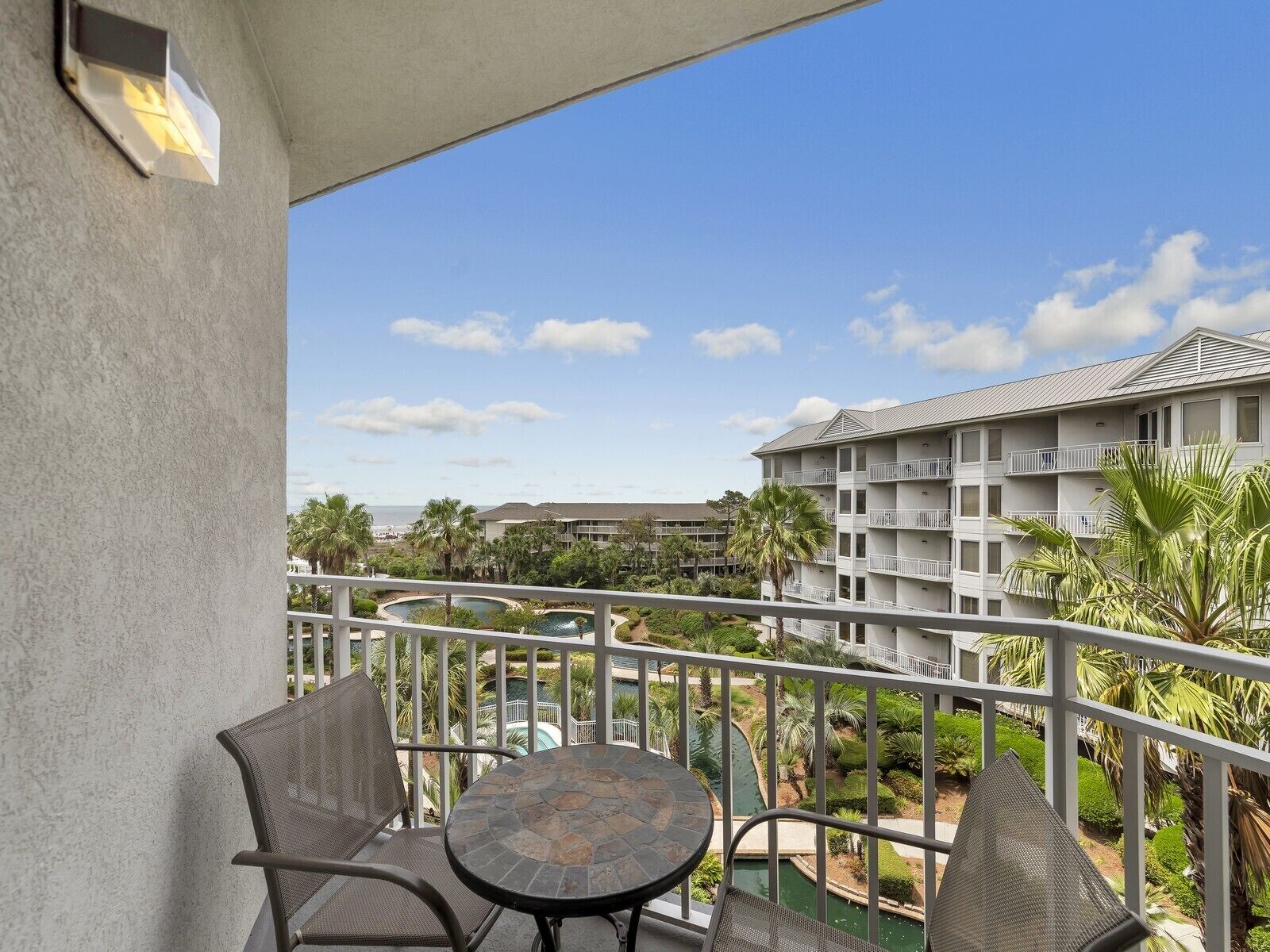 A balcony with two wicker chairs and a stone table, overlooking a pool and a building with multiple balconies.