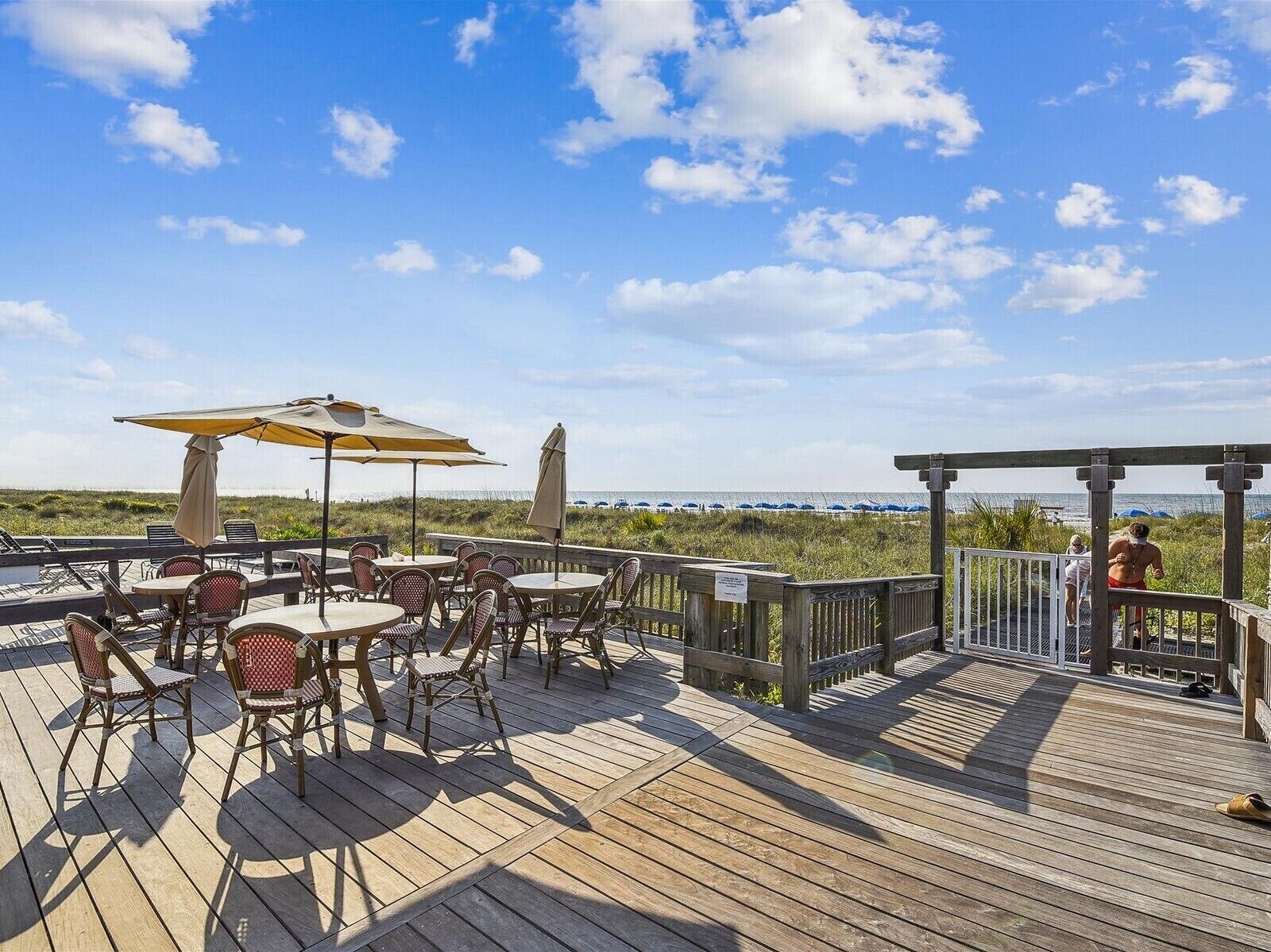 A wooden deck with outdoor seating, including tables and chairs, overlooking a beach with umbrellas and a wooden structure.