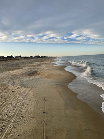 A sandy beach with footprints and tire tracks, a calm sea, and a clear sky with clouds.