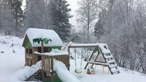 A wooden playhouse with a slide and swing set, surrounded by snow-covered trees.