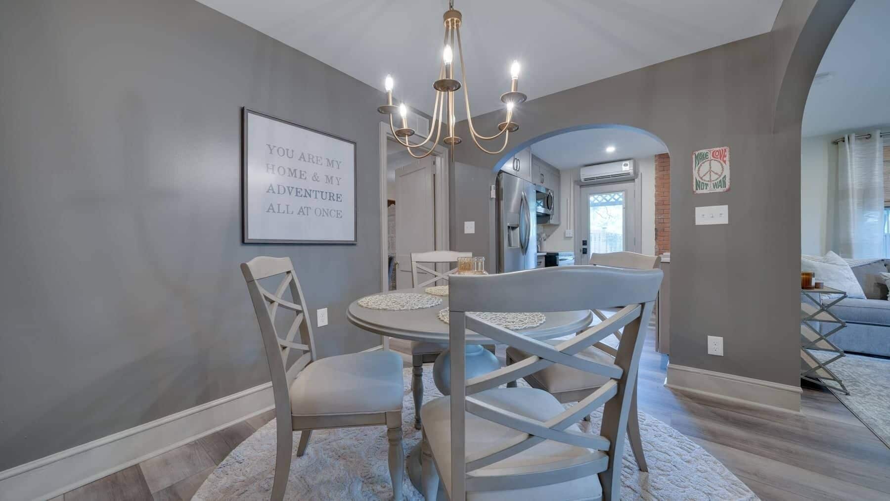 A dining area with a round table, four chairs, a chandelier, and a framed inspirational quote on the wall.