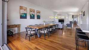 Un vaste espace de salle à manger avec une longue table en bois et des chaises modernes, un canapé blanc et une cuisine en arrière-plan.